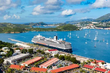 View of a large cruise ship docked at a vibrant coastal city, surrounded by colorful buildings and boats floating in the blue sea, with rolling hills and islands in the distance.